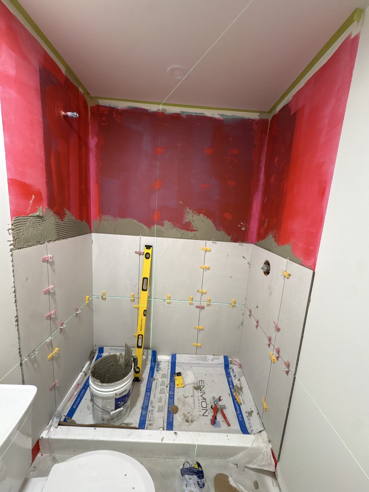 Tile going in over a liquid waterproofing membrane, checked with laser and level — the finish stage, fitted inside the building's allowed work hours